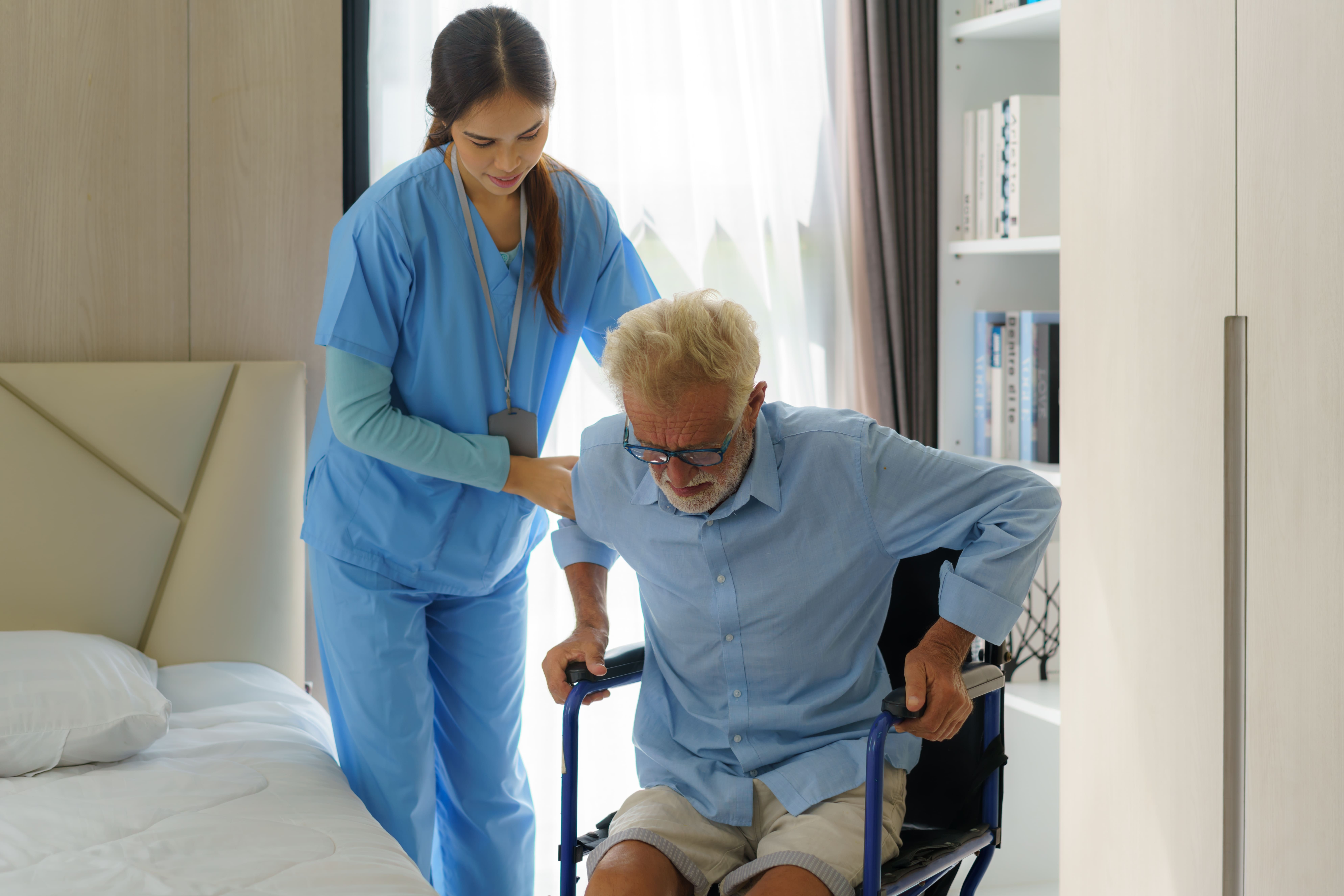 A woman carer helps an older man out of his wheelchair in his bedroom