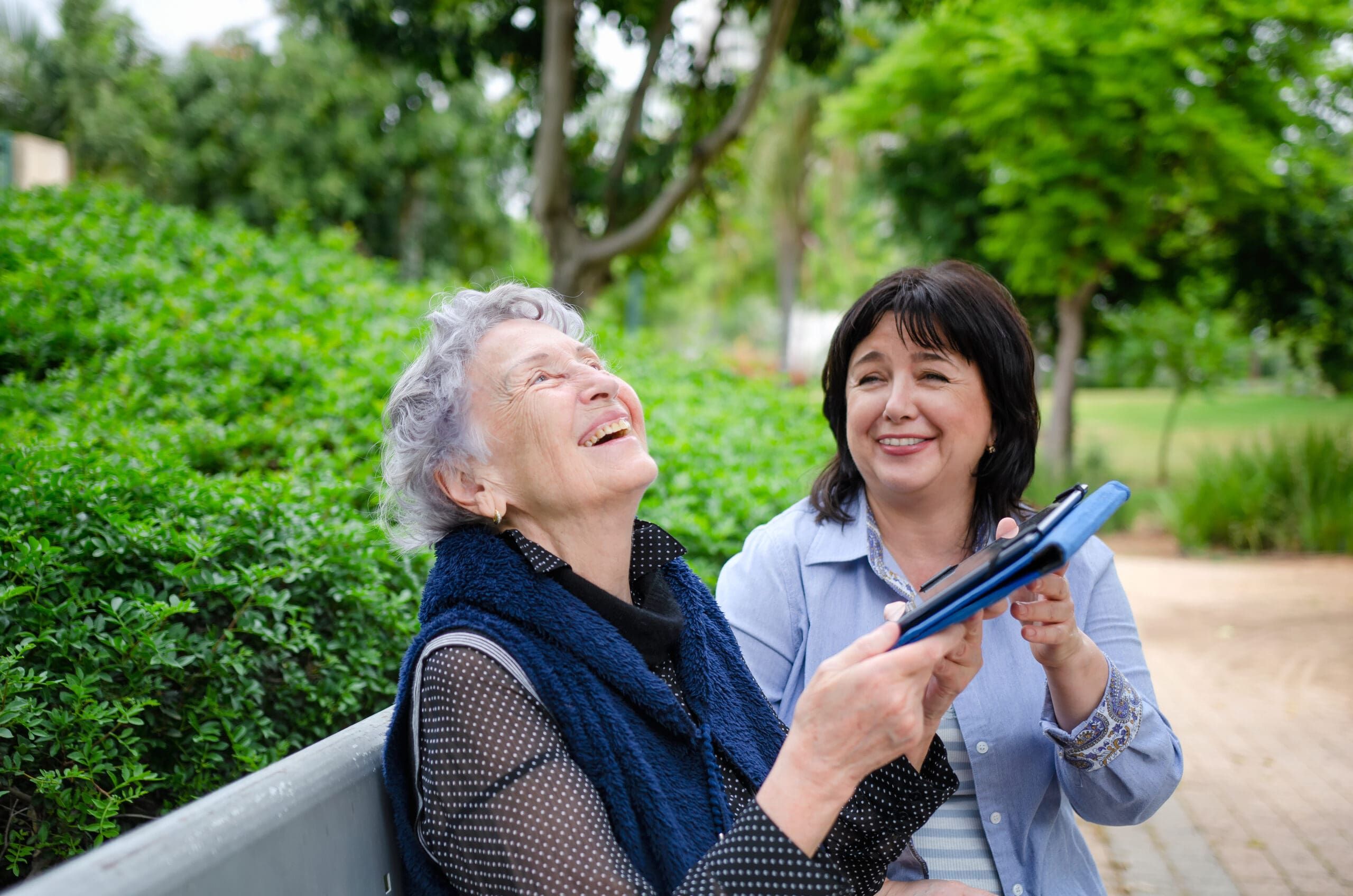 A woman and her carer share a laugh and a joke at an electronic tablet on a bench outdoors