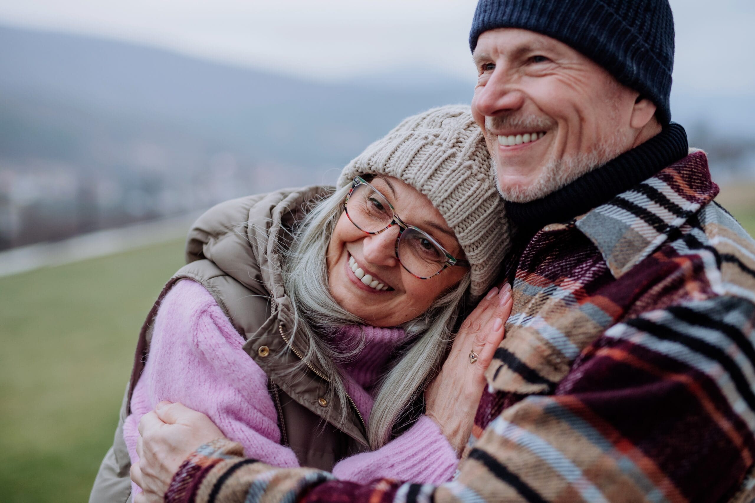 An older couple embrace in hats and scarves as they look on at the view on a walk outdoors