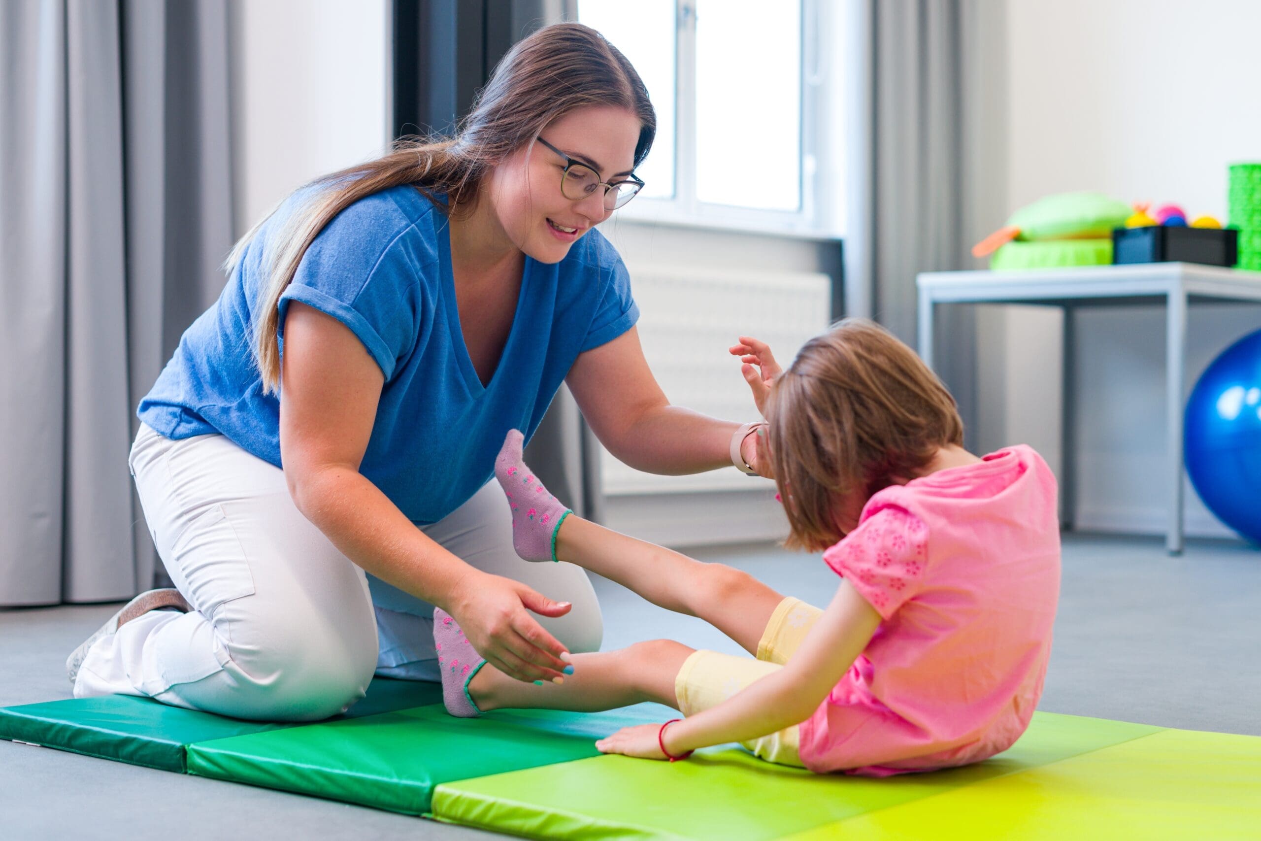 A child with a disability does exercises with her carer in a physical therapy session on some colourful mats