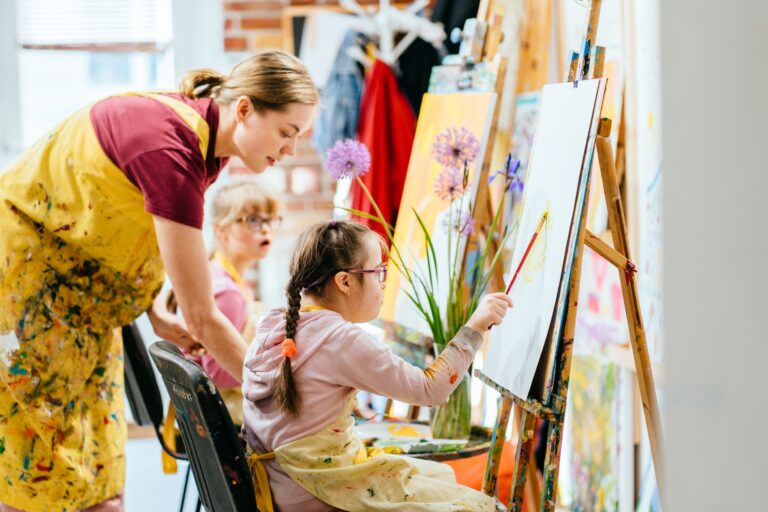A young girl sits at an easel painting fluffy pink flowers while her art teacher gives helpful feedback